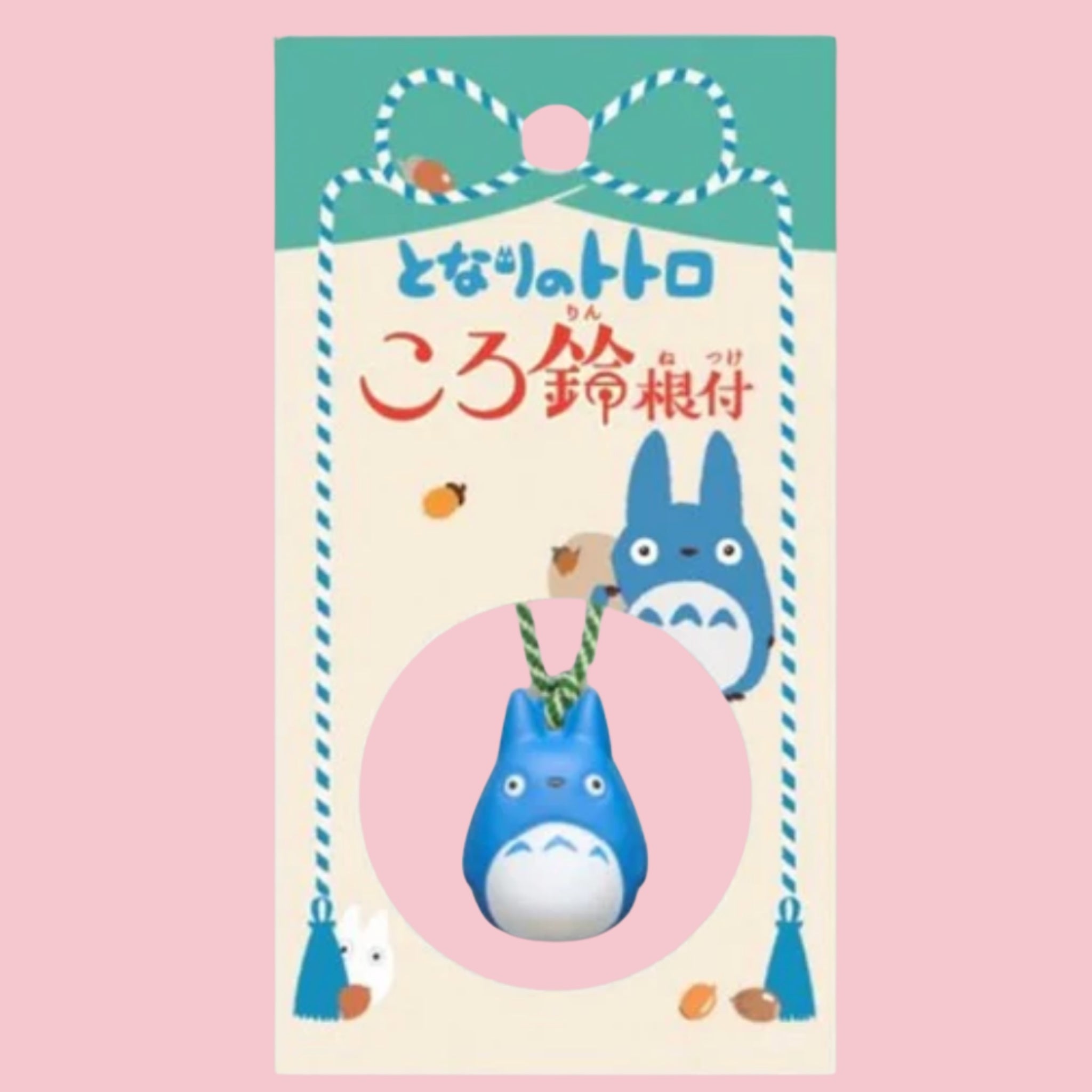 Original Japanese packaging card for the My Neighbor Totoro bell charm, featuring the character illustration and Japanese title in red text on a green and cream backdrop, with the medium blue Totoro bell charm shown hanging on its braided cord inside a circular pink window at the bottom of the card.