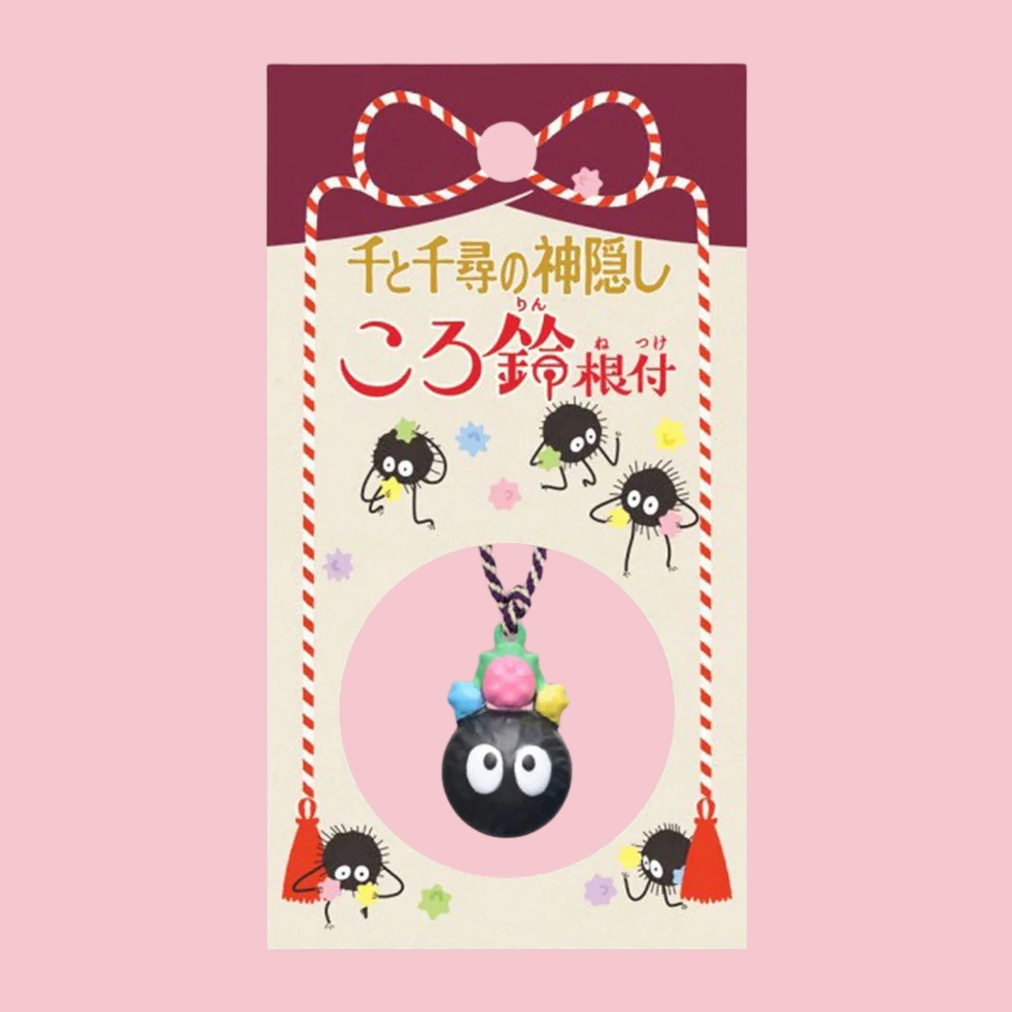Original Japanese packaging card for the Spirited Away Soot Sprite bell charm, featuring several soot sprite illustrations and Japanese title in gold and red text on a maroon backdrop, with the round black soot sprite bell charm topped with colorful candy shown hanging on its cord inside a circular pink window at the bottom of the card.