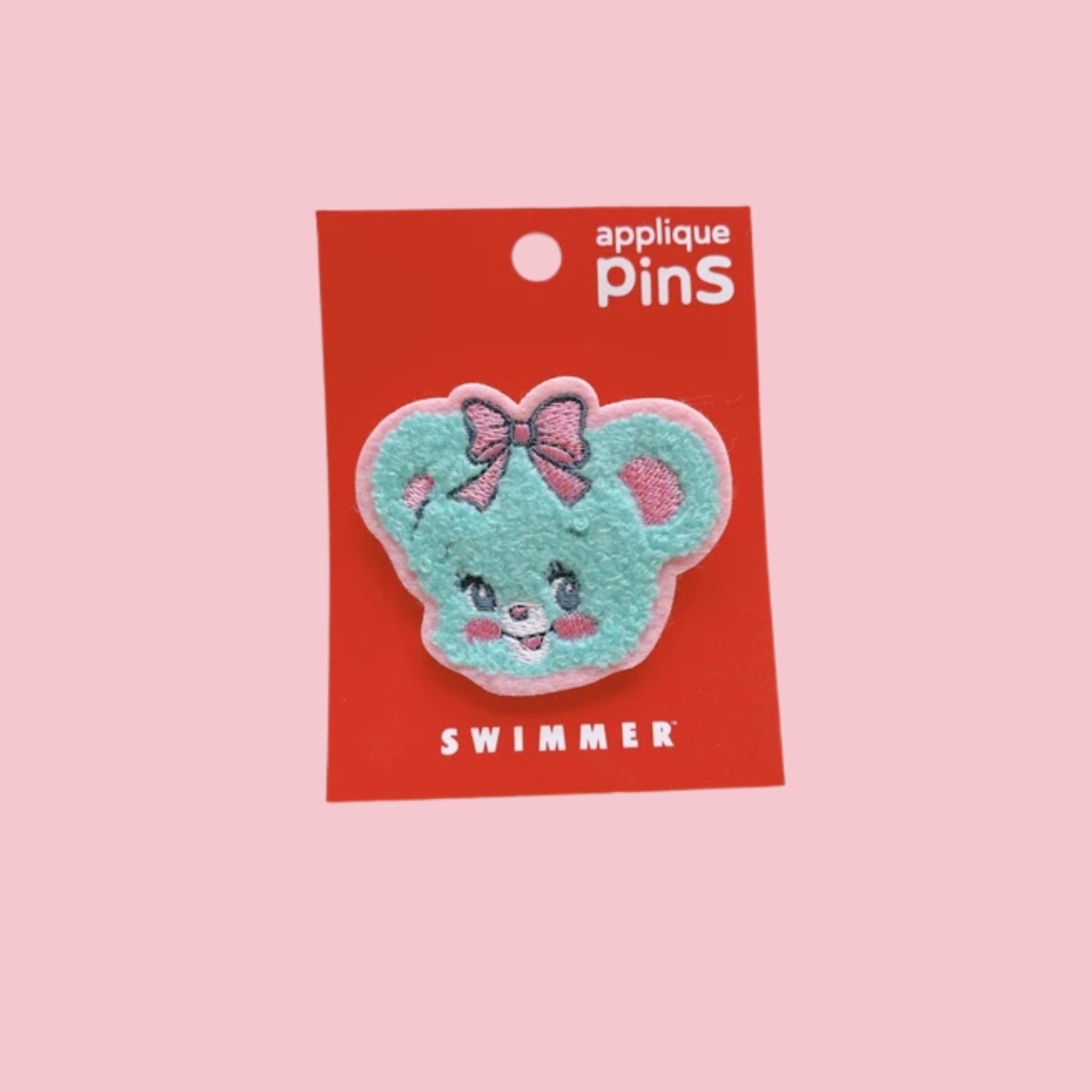 Teal fuzzy bear face applique pin with a pink bow, attached to a red Swimmer card on a pink background.