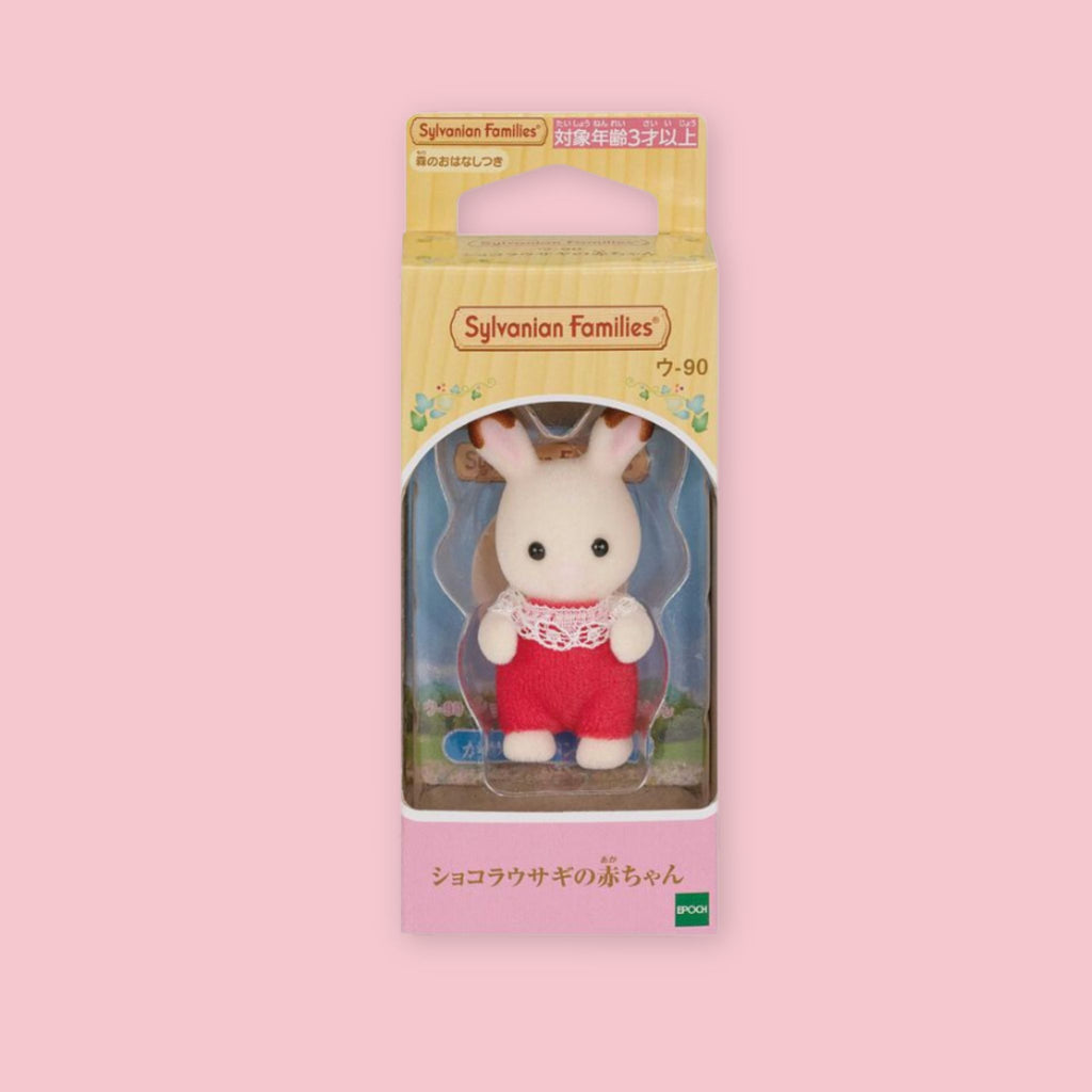 Baby Sylvanian Families chocolate rabbit figure in original Japanese box packaging, wearing a red outfit with white lace collar, photographed against a pastel pink background.