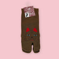 Brown Japanese tabi socks with a cat face design, shown in package against a pink background.