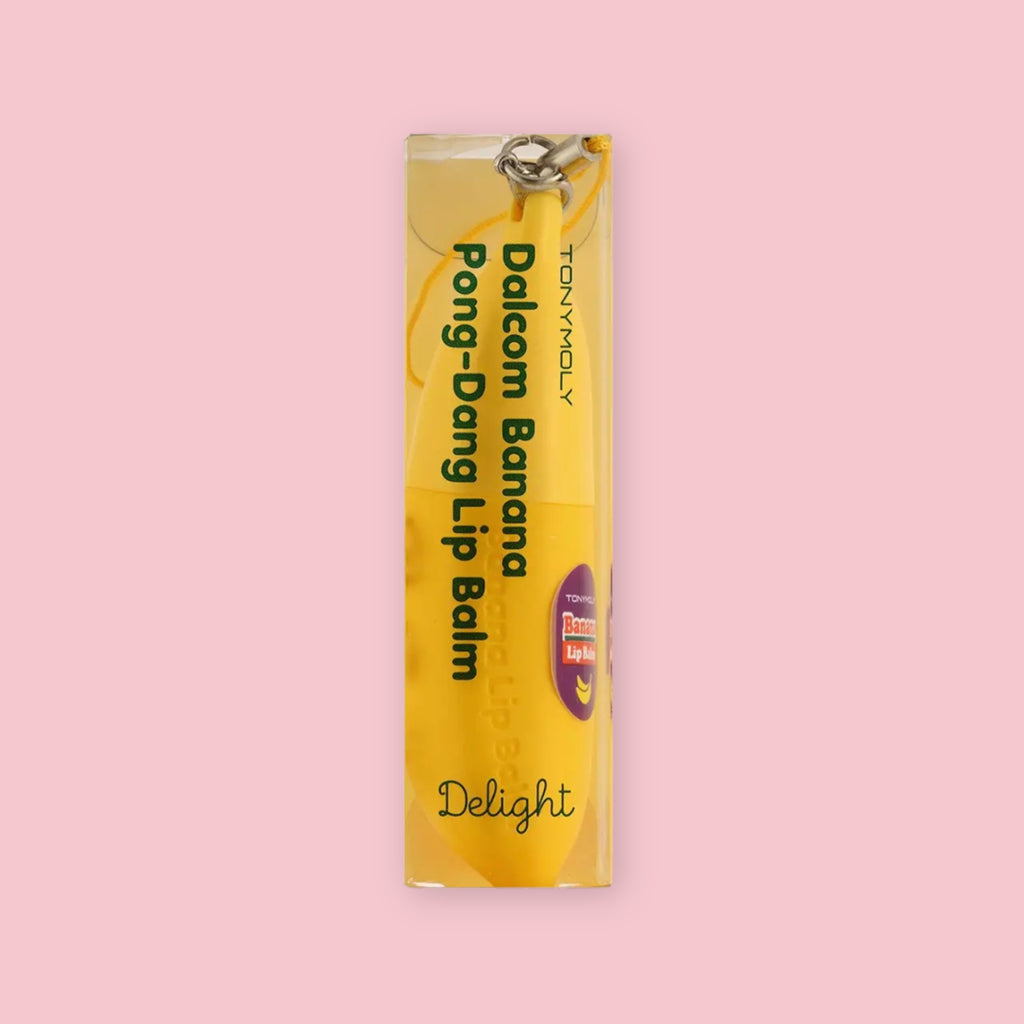 TonyMoly Banana Lip Balm shown inside its clear plastic packaging printed with "Dalcom Banana Pong-Dang Lip Balm" and "Delight," on a pink background.