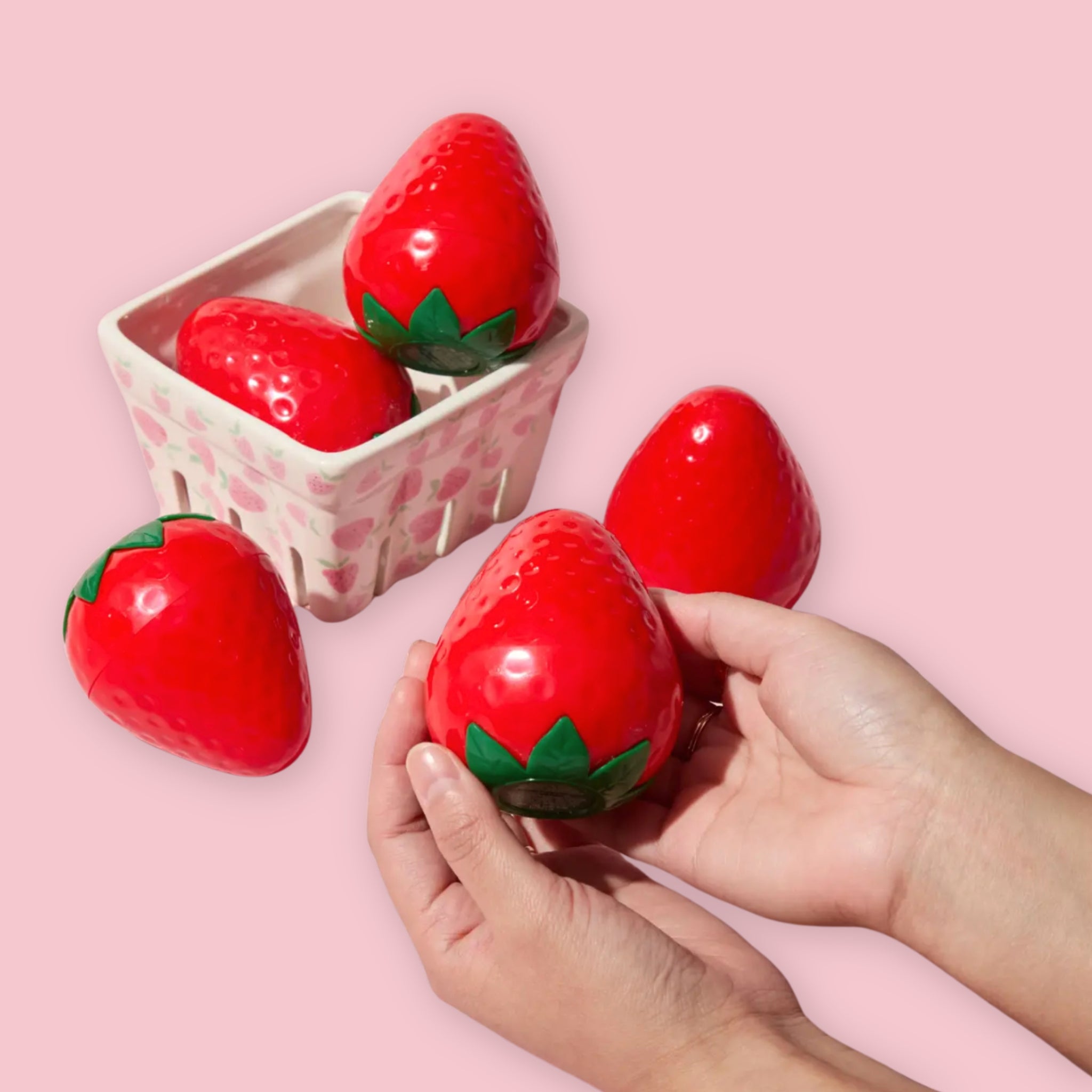 TonyMoly strawberry-shaped hand cream containers held in hands and arranged in a small basket, on a pink background.