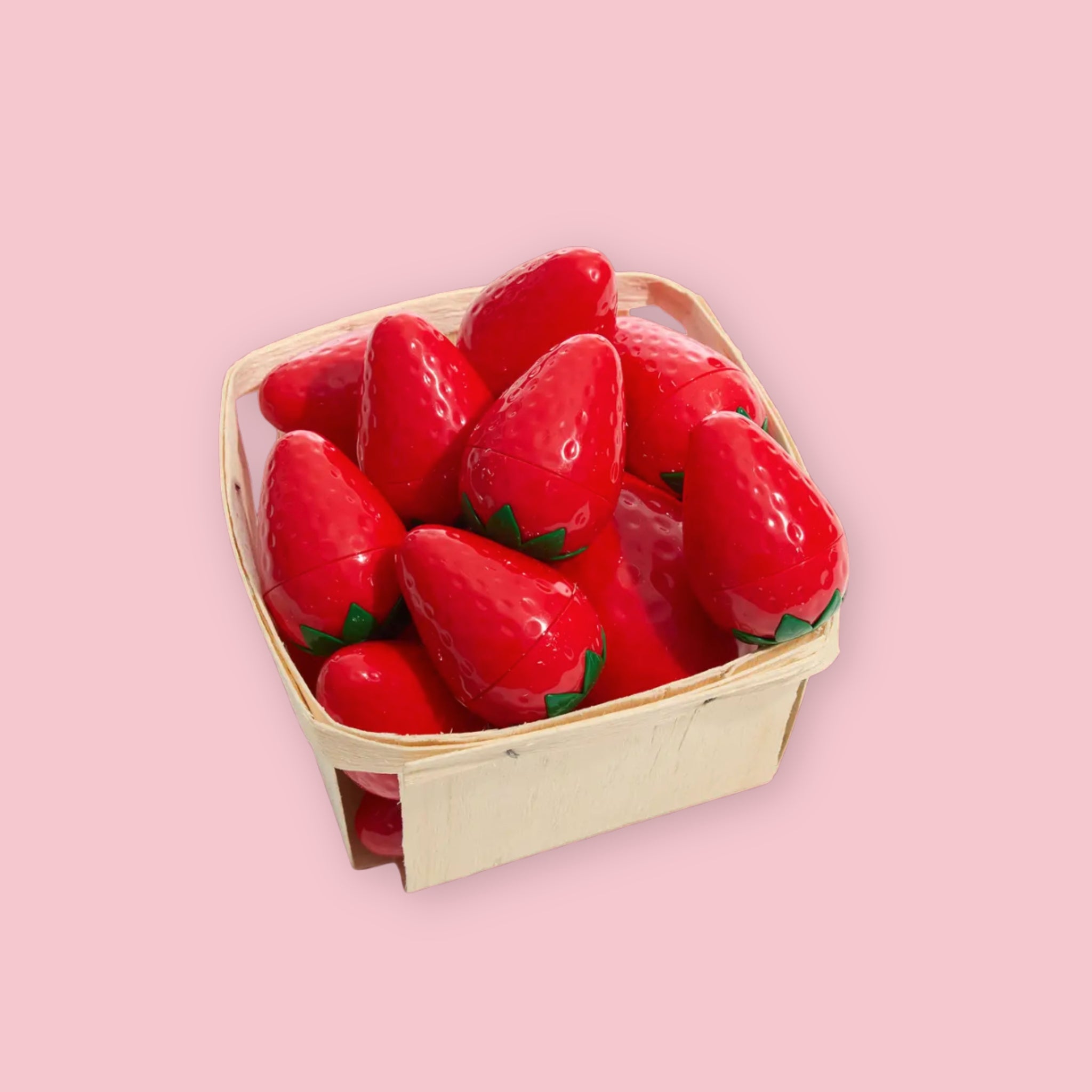 Multiple TonyMoly Strawberry Lip Balms shaped like whole strawberries, piled together in a small wooden berry basket, on a pink background.