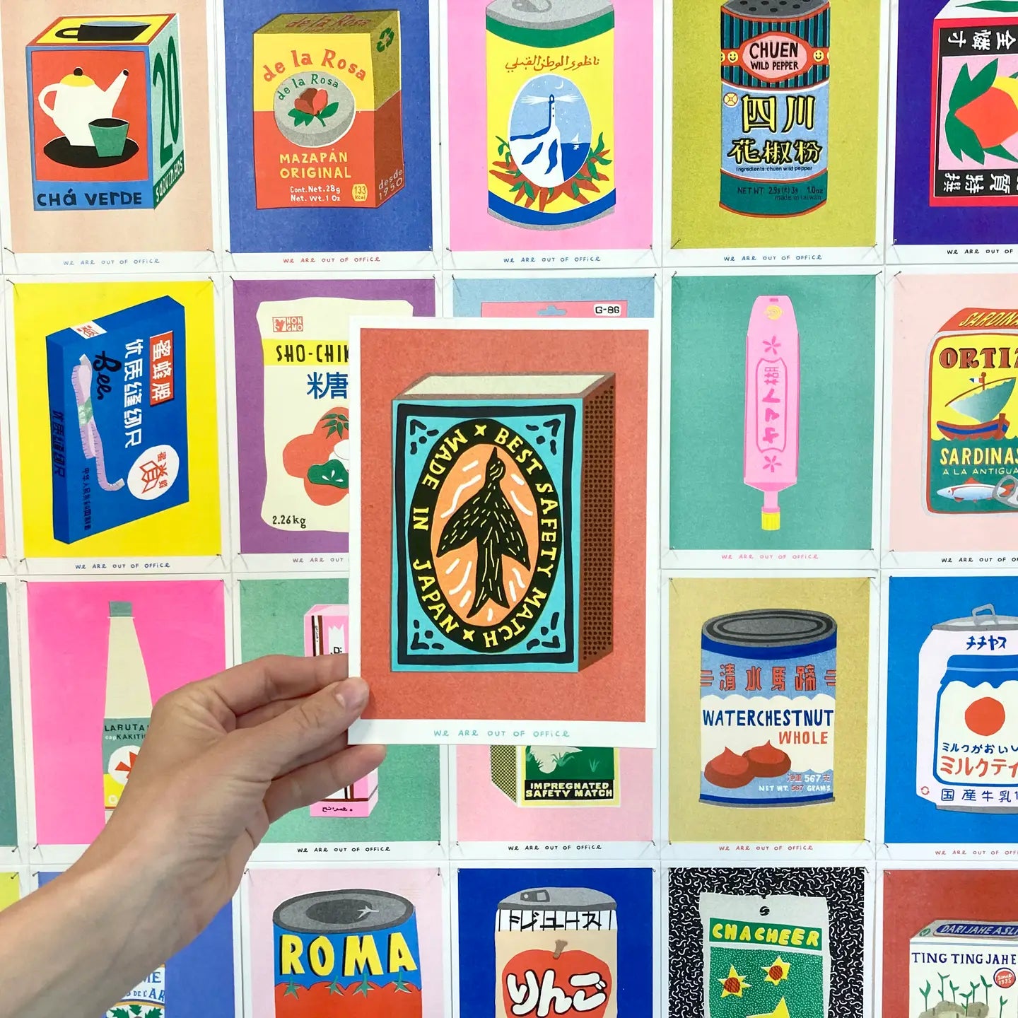 Hand holding a We Are Out of Office art print featuring a vintage Japanese safety-match design, displayed against a wall of colorful packaging-inspired prints.