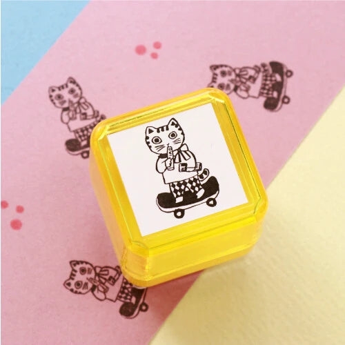 Yumi Kitagishi self-inking stamp in yellow with a Cat Going Out design showing a cat on a skateboard, with black stamp impressions visible on paper in the background, on a pink and yellow surface.