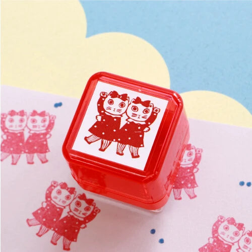 Yumi Kitagishi self-inking stamp in red with a Duo design showing two cat girls in matching polka dot dresses, with red stamp impressions visible on paper in the background, on a yellow and blue surface.