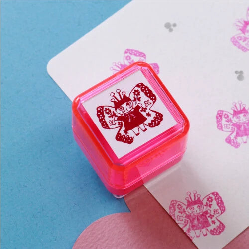 Yumi Kitagishi self-inking stamp in pink with a Fairy design showing a cat girl with butterfly wings and a crown, with pink stamp impressions visible on paper in the background, on a blue surface.